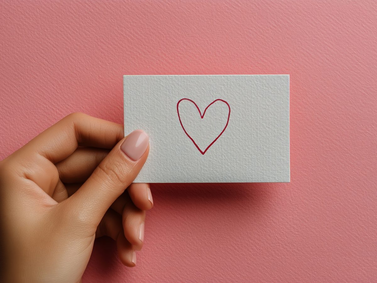 A hand gracefully holds a white card featuring a simple red heart sketch, set against a soft pink backdrop, symbolizing love and warmth in a creative expression.